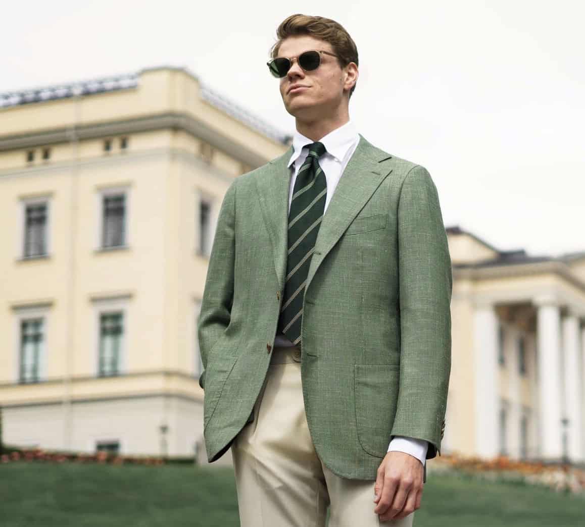 Man wearing sunglasses, green blazer and trousers standing in front of a colonial building