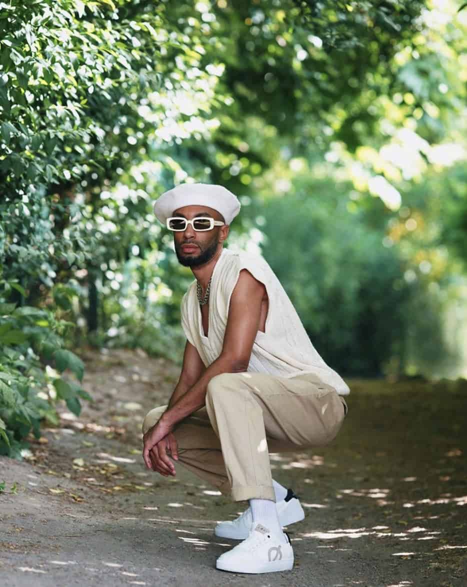 man squatting down wearing a beret, white glasses and a sweater vest