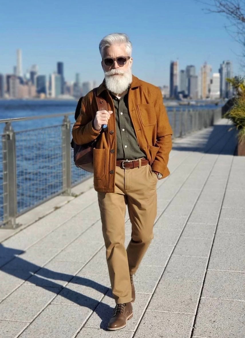 man walking on a boardwalk holding one strap of a backpack, groomed white beard and sunglasses