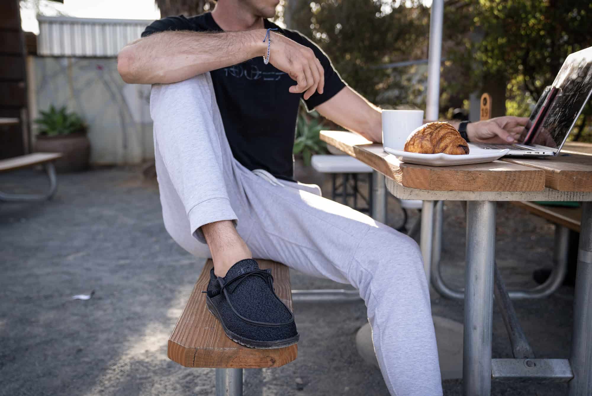 man sitting on bench, wearing hey dude shoes, looking at laptop