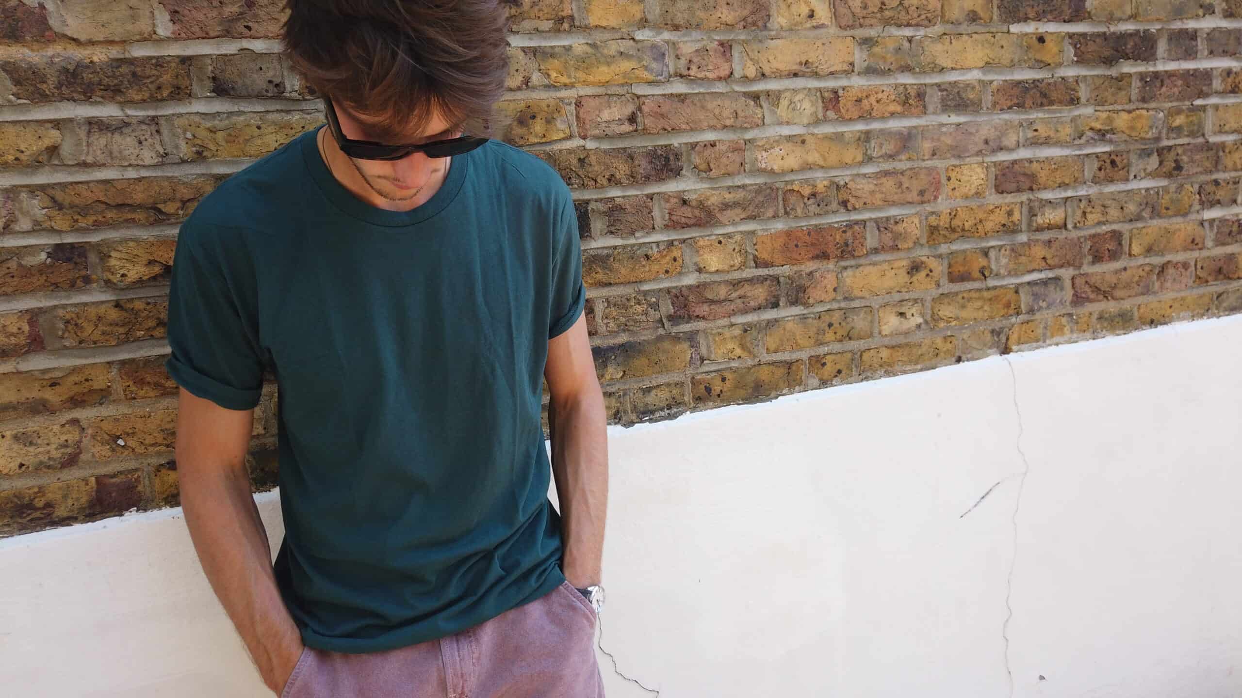 man looking down standing against a brick wall wearing a green true classic tee
