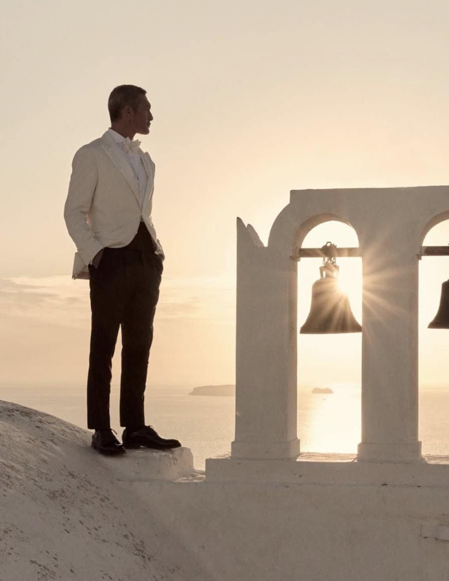 man in a suit standing on a cliff in front of a bell tower at sunset