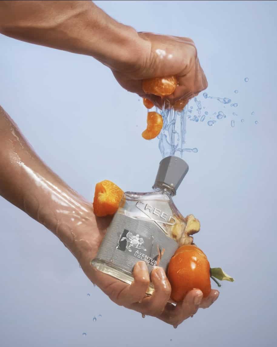 A bottle of Creed Cologne with hands squeezing oranges over the bottle
