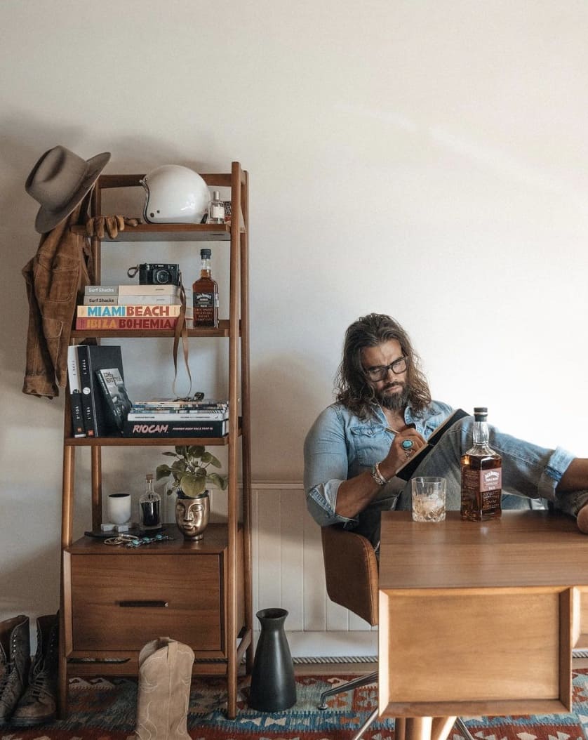 man sitting at a desk with his feet up writing in a book, wearing denim with whiskey on the table