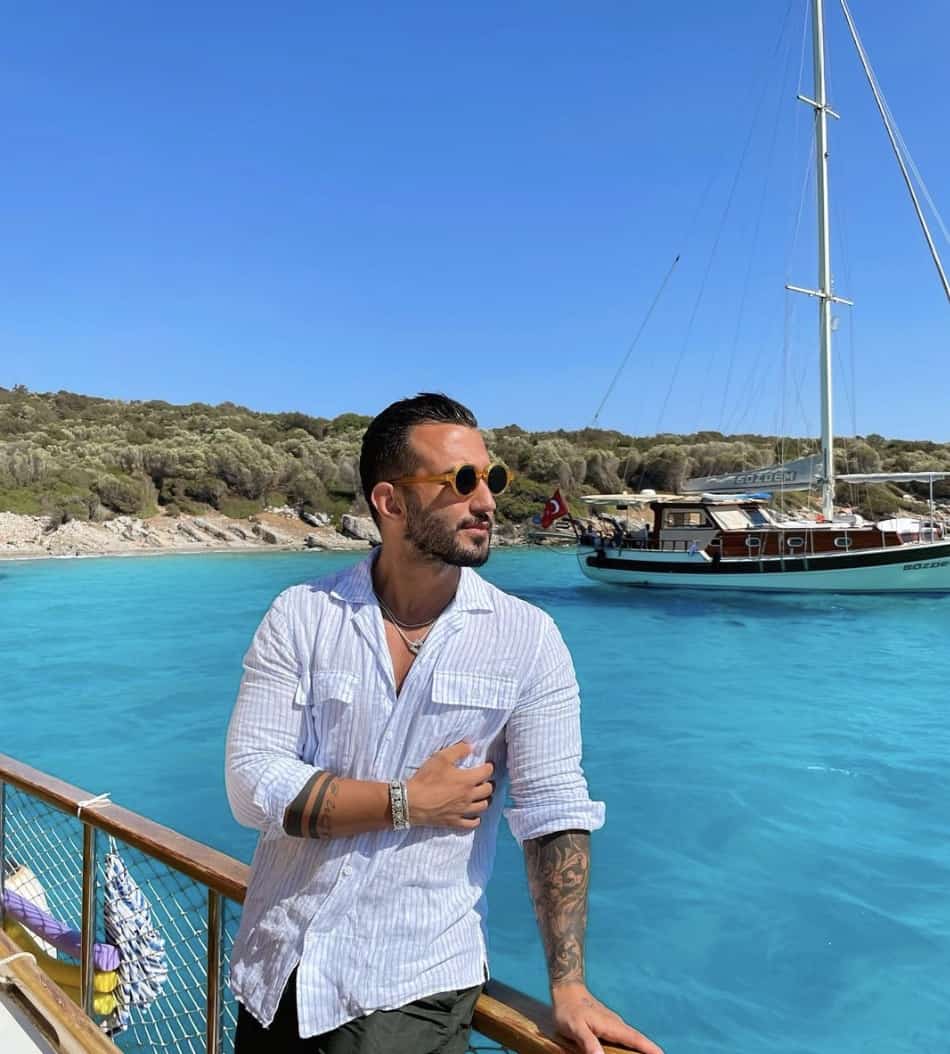 man standing in front on a sail boat with very blue water behind him, smiling looking away from the camera