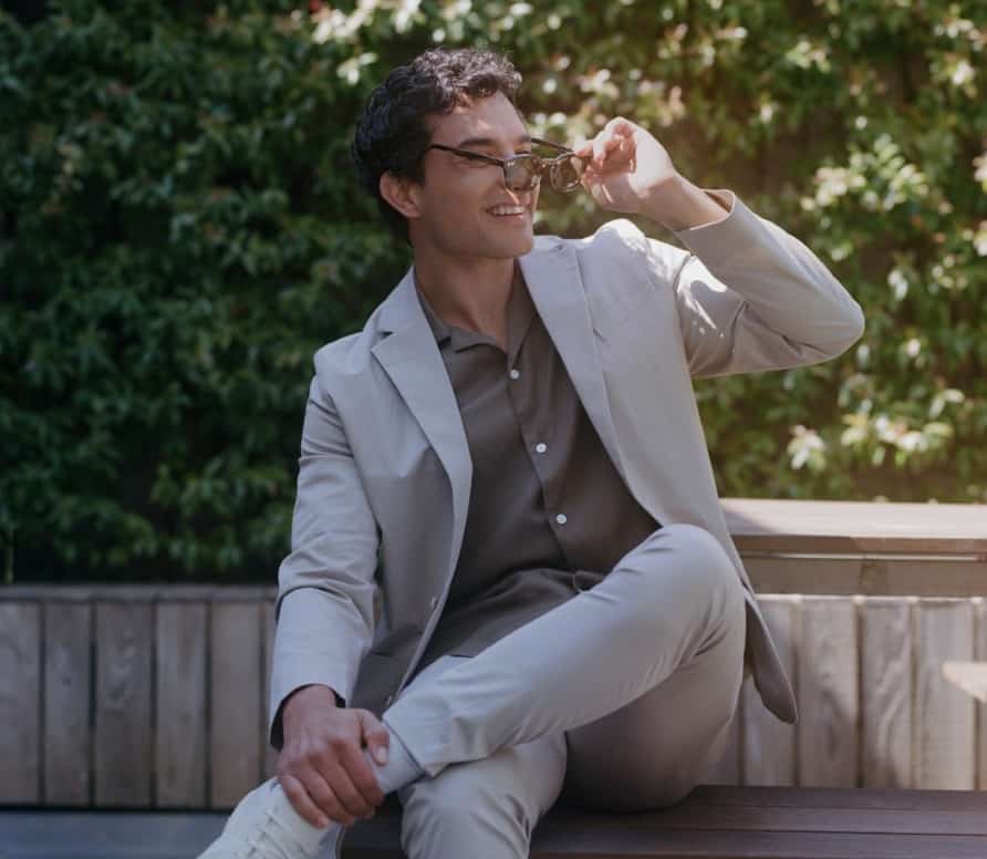 man sitting on a bench outside, crossed legs, in a light colored suit and putting sunglasses on 