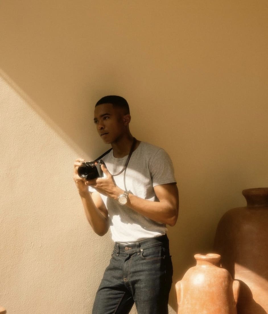 man in a slim fit t-shirt, holding a camera, leaning in a shadow against a yellow wall 
