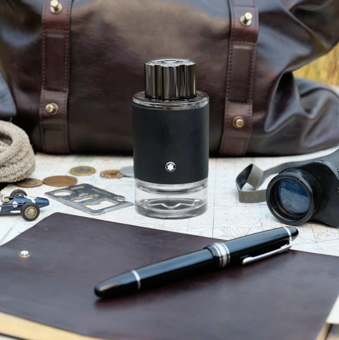 Montblanc cologne bottle on a desk with a leather weekender bag behind it, notebook, pen scattered around