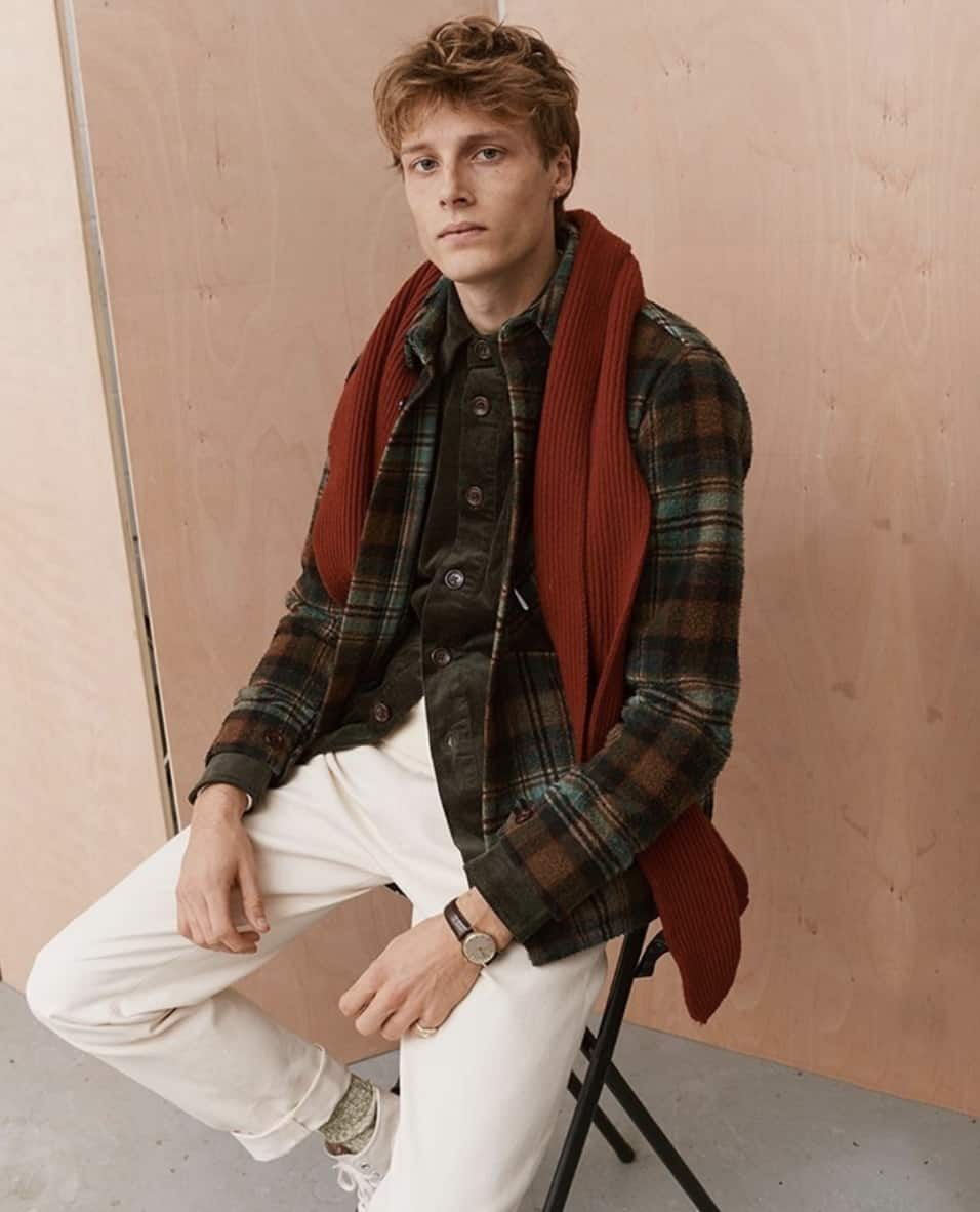 Man sitting on a chair, looking at the camera, wearing white pants, a green flannel and an orange scarf. 