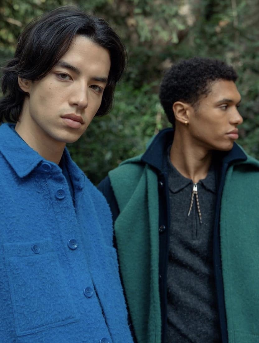 two men outside, one looking at the camera in a blue shirt, one looking away in a green shirt.