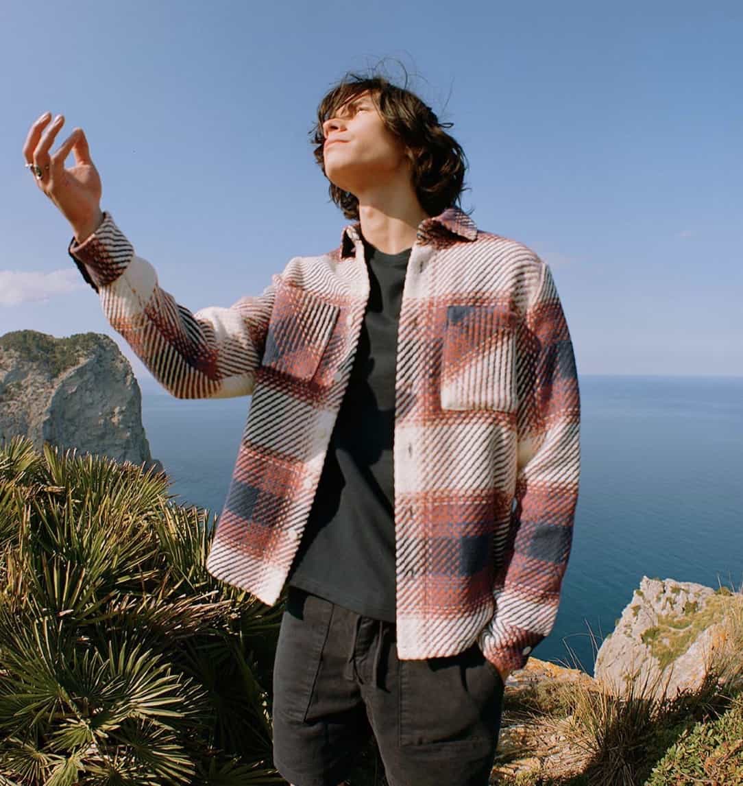 man holding his arm in the air, cliff-side with the ocean behind him. Wearing a red flannel, black t-shirt and black shorts. 