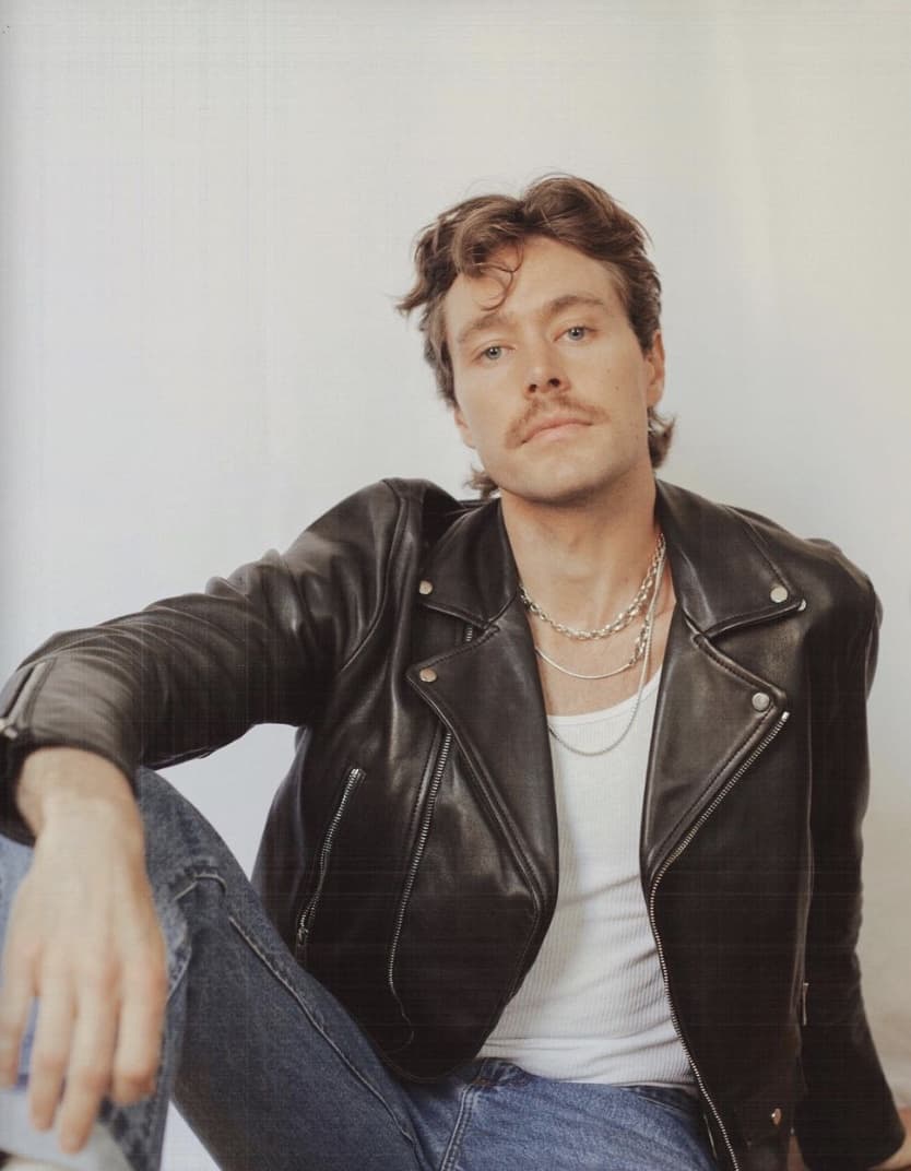 man sitting looking at the camera, wearing a leather jacket, tank top and jeans