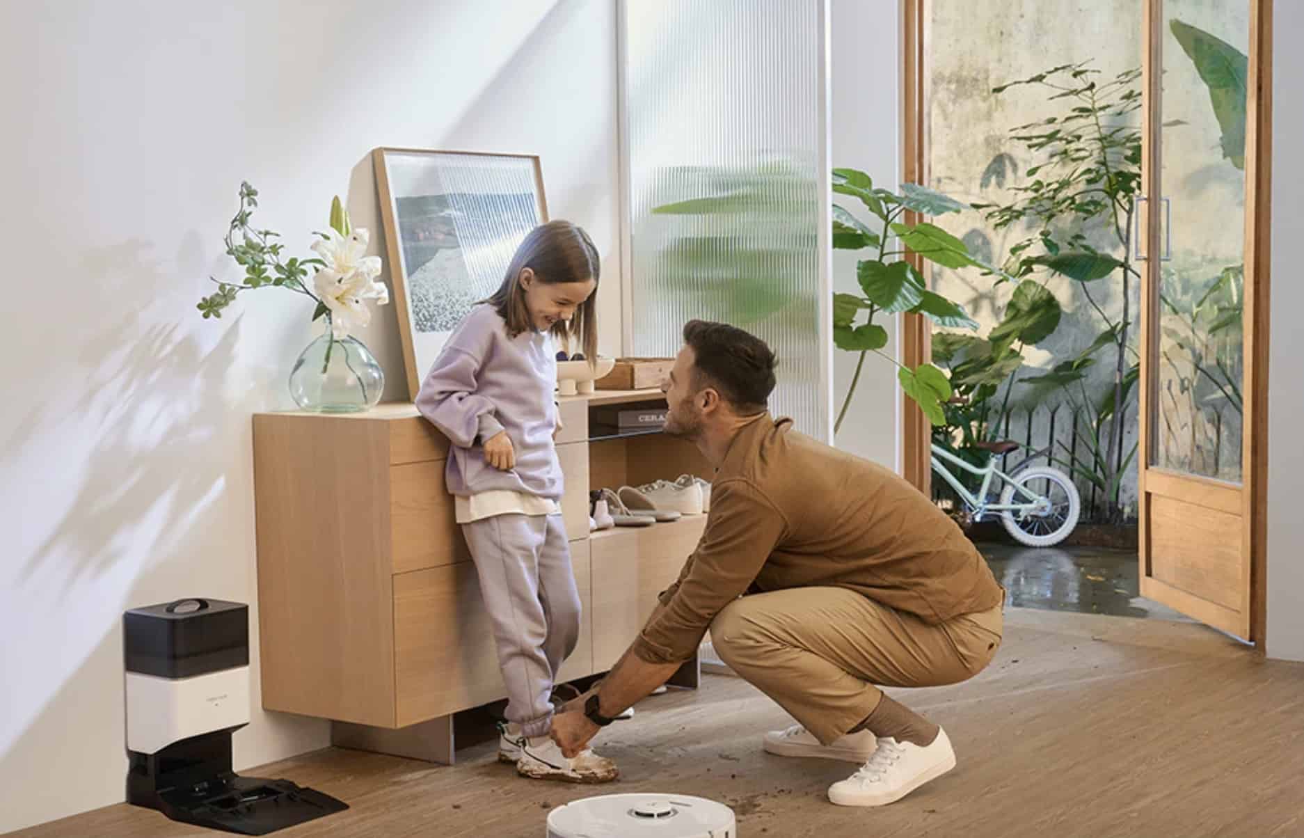 man helping a child take off muddy shoes while robot vacuum cleans around them