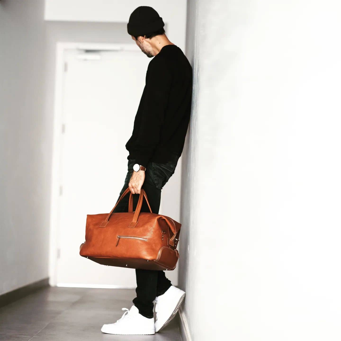 man carrying a leather weekender while leaning on the wall