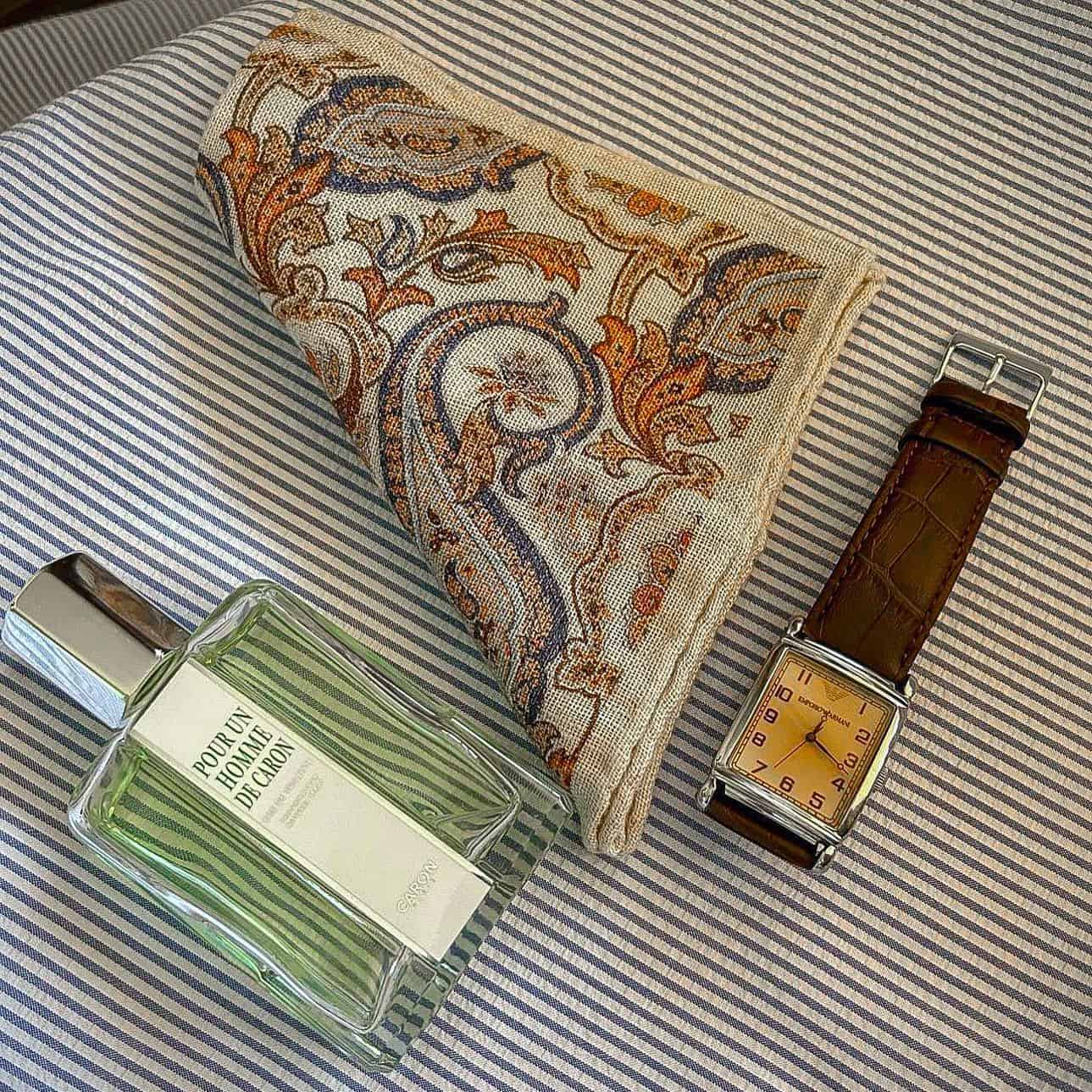 a printed handkerchief in-between a bottle of cologne and a watch