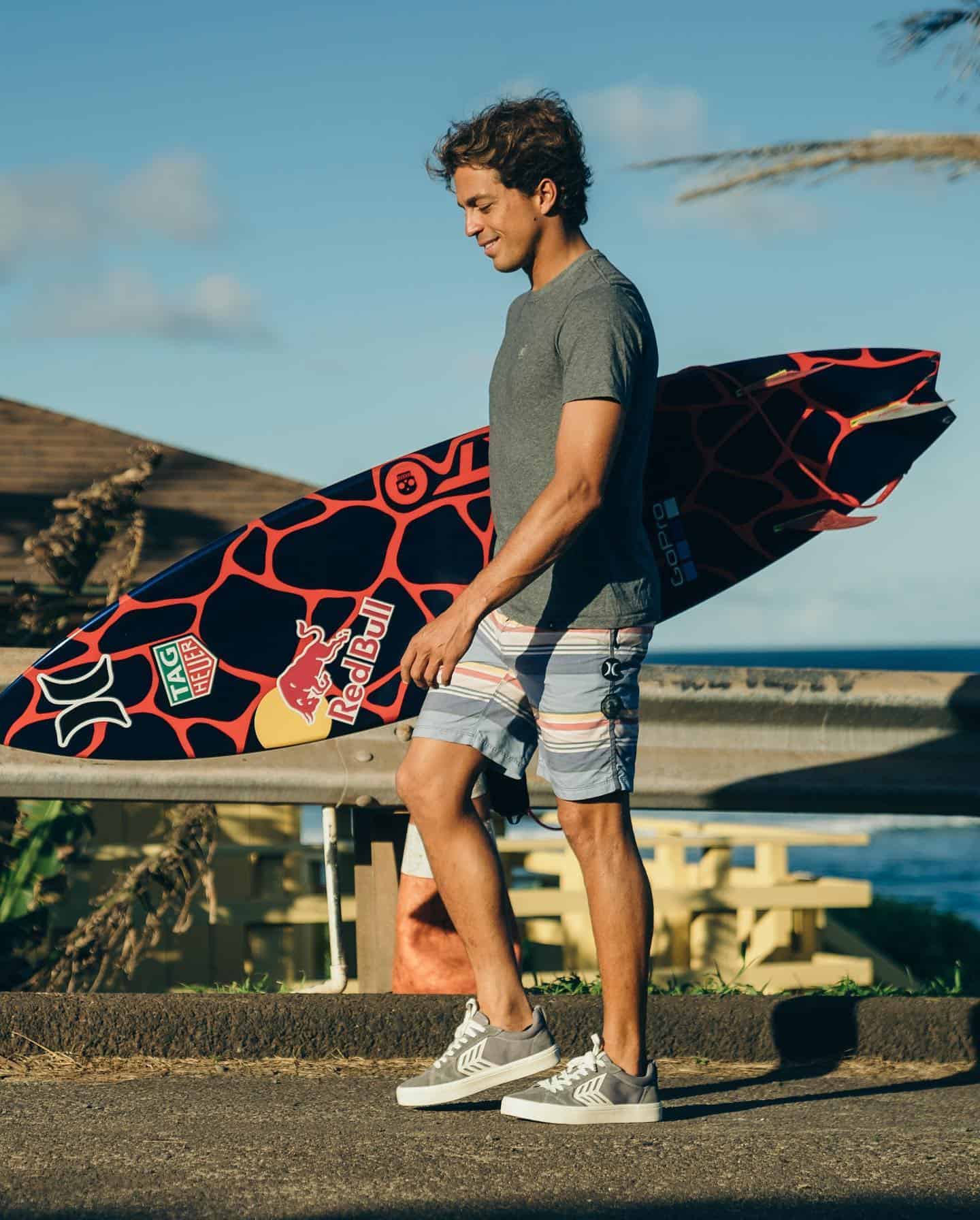 man carrying a surfboard wearing a gray shirt and shorts with stripe