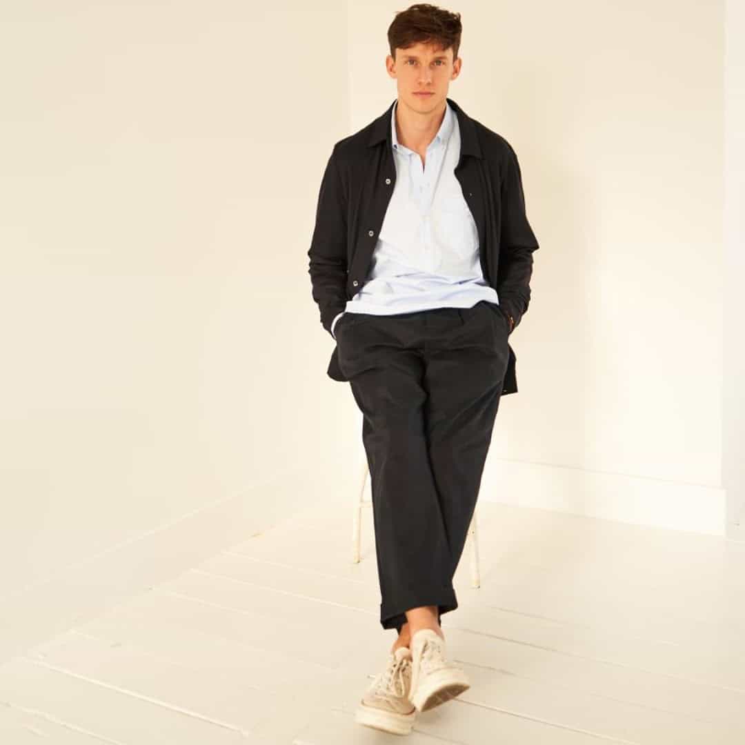 man sitting on a stool wearing black jacket over a white shirt and black pants