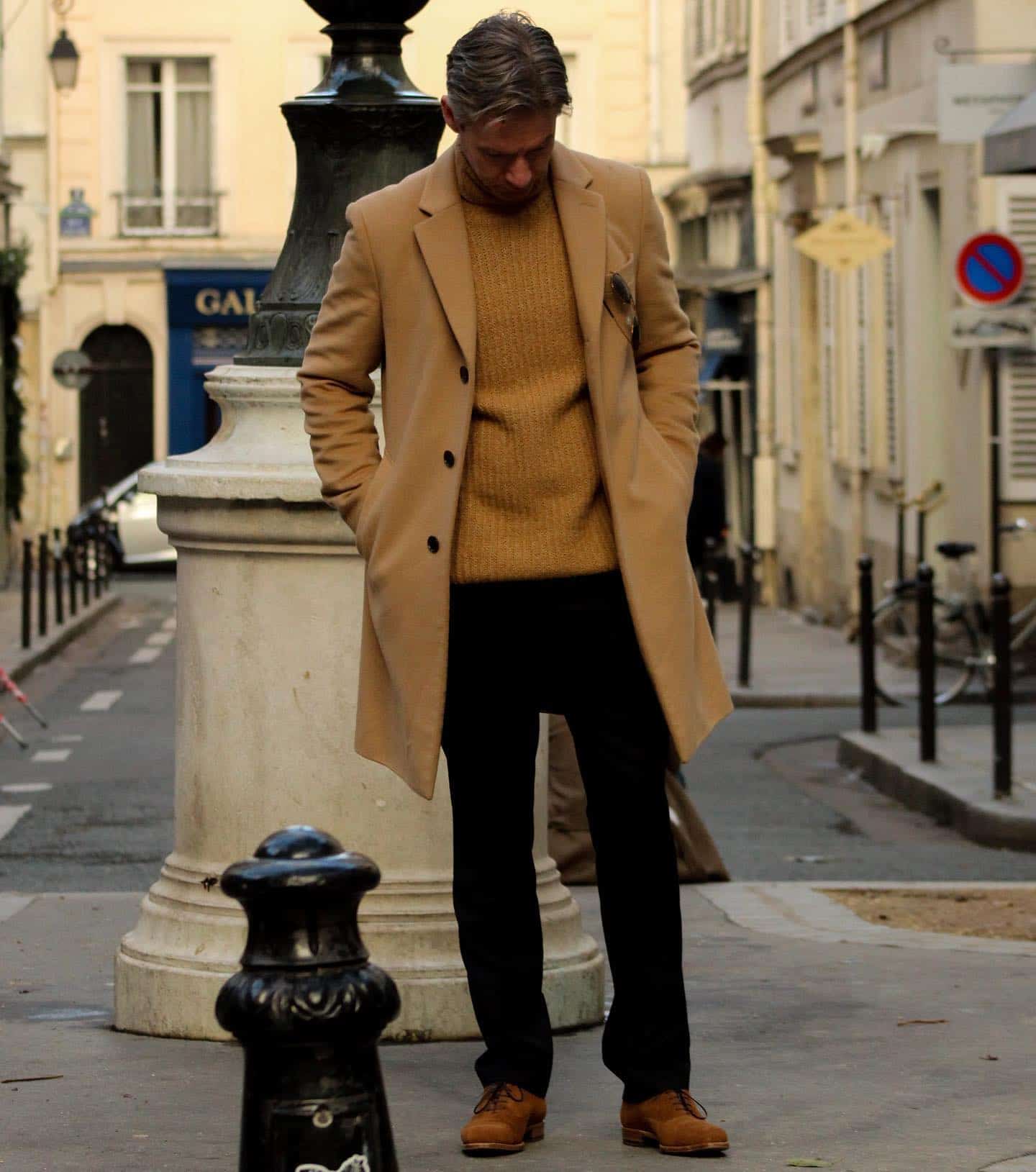 man in a long brown coat checking his shoes