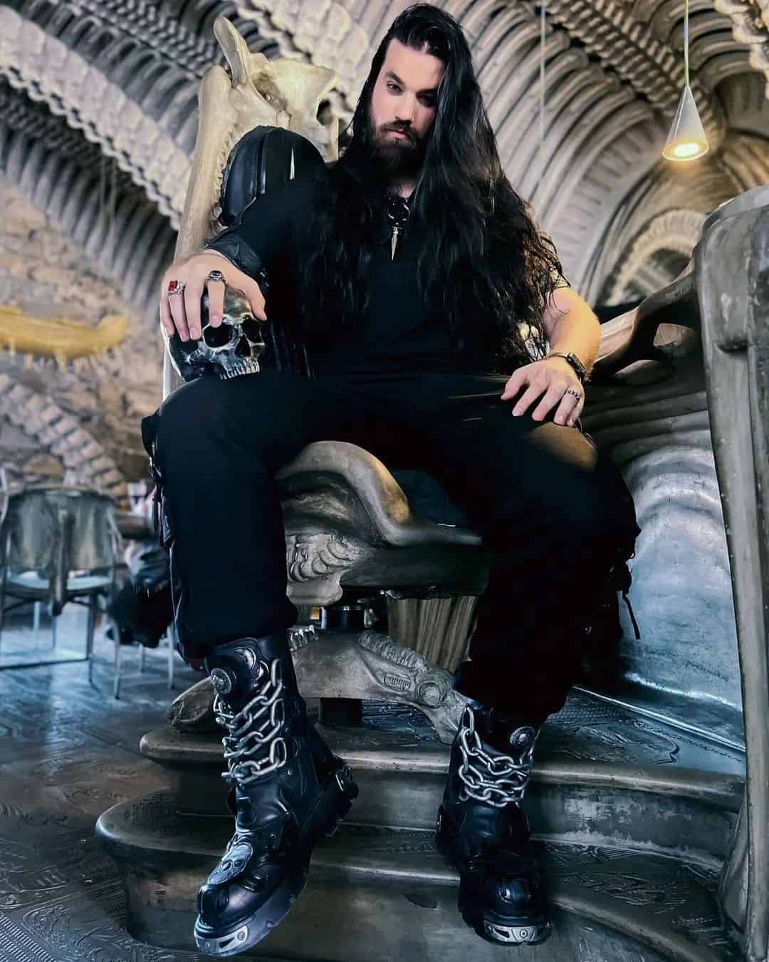 man with long hair sitting on a chair wearing an all black outfit