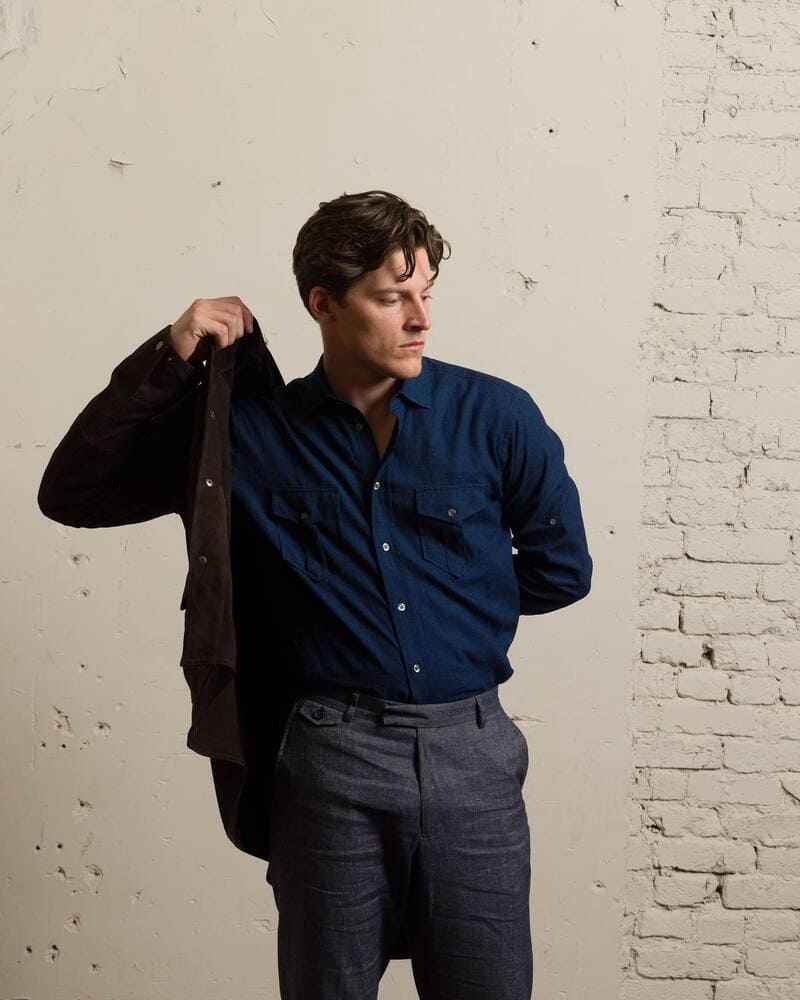 man wearing a blue shirt and grey pants putting on a brown jacket