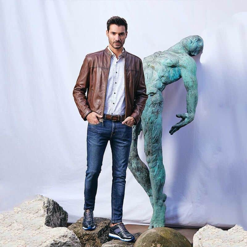 man in brown leather jacket and blue jeans standing infront of a statue