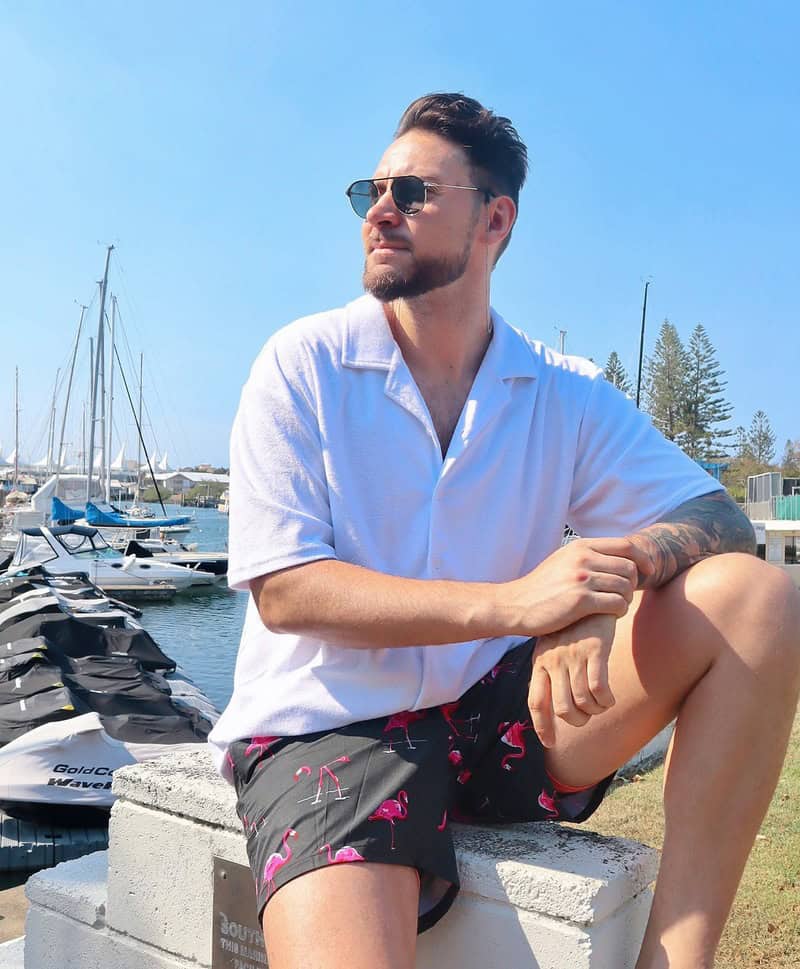 man sitting by the docks wearing white shirt and black shorts with flamingo print