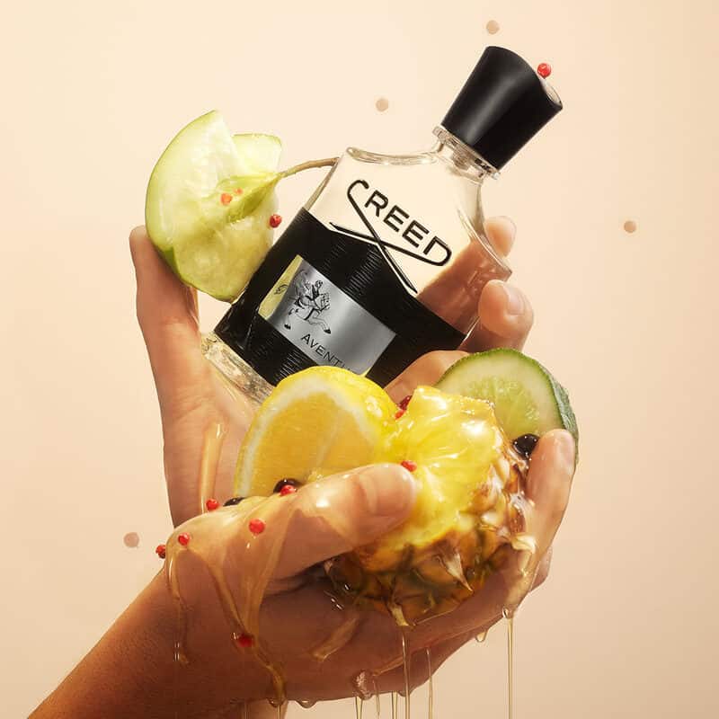 A man holding with both hands a bottle of Creed Aventus cologne with slices of pineapple, lemon lime and half eaten pear