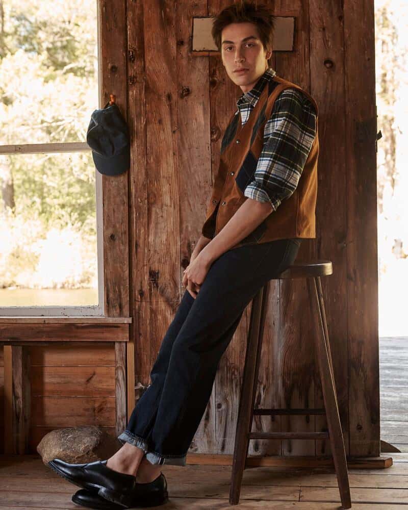 man sitting on a wooden stool wearing a checked shirt layered with a brown vest