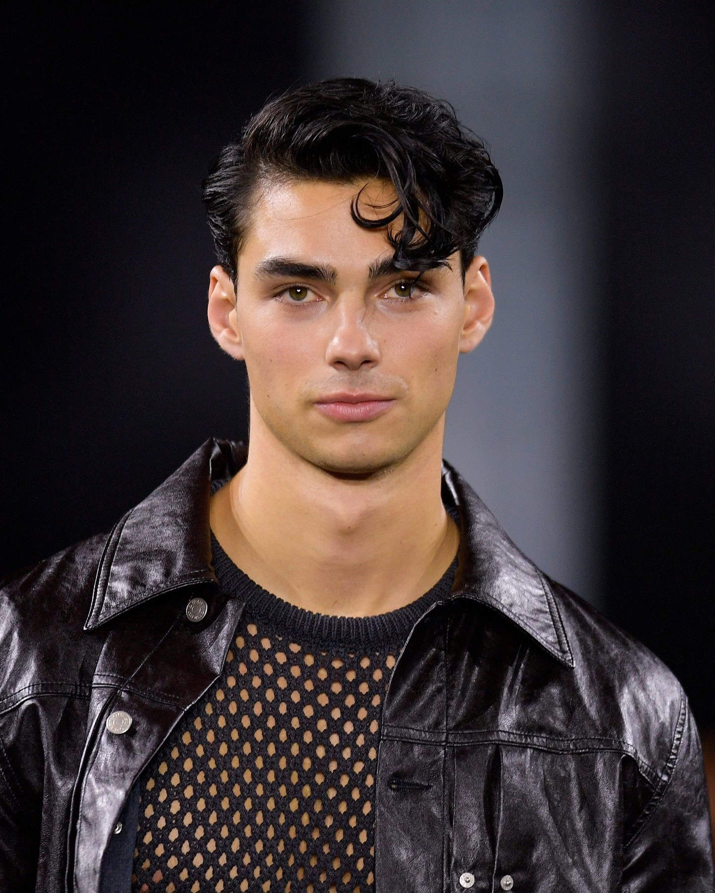 man with black wavy hair wearing a leather jacket over a mesh t-shirt