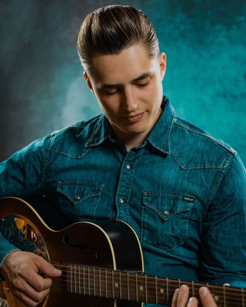 man with a pompadour hairstyle wearing a denim shirt while playing a guitar
