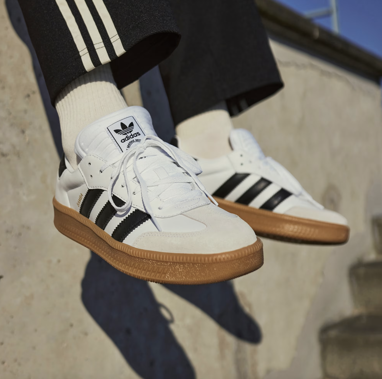 White Adidas Samba sneakers with black stripes on gum sole, paired with beige socks and black pants, sitting outdoors with a concrete background. Perfect casual footwear for a stylish everyday look.
