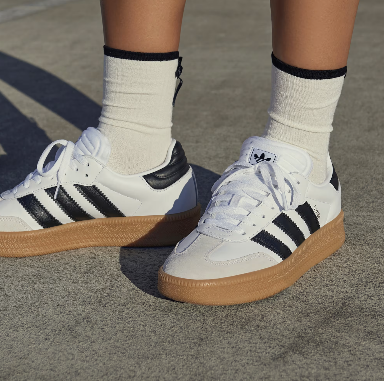 Adidas shoes with black stripes and tan soles on a person's feet, paired with white crew socks on a concrete surface.