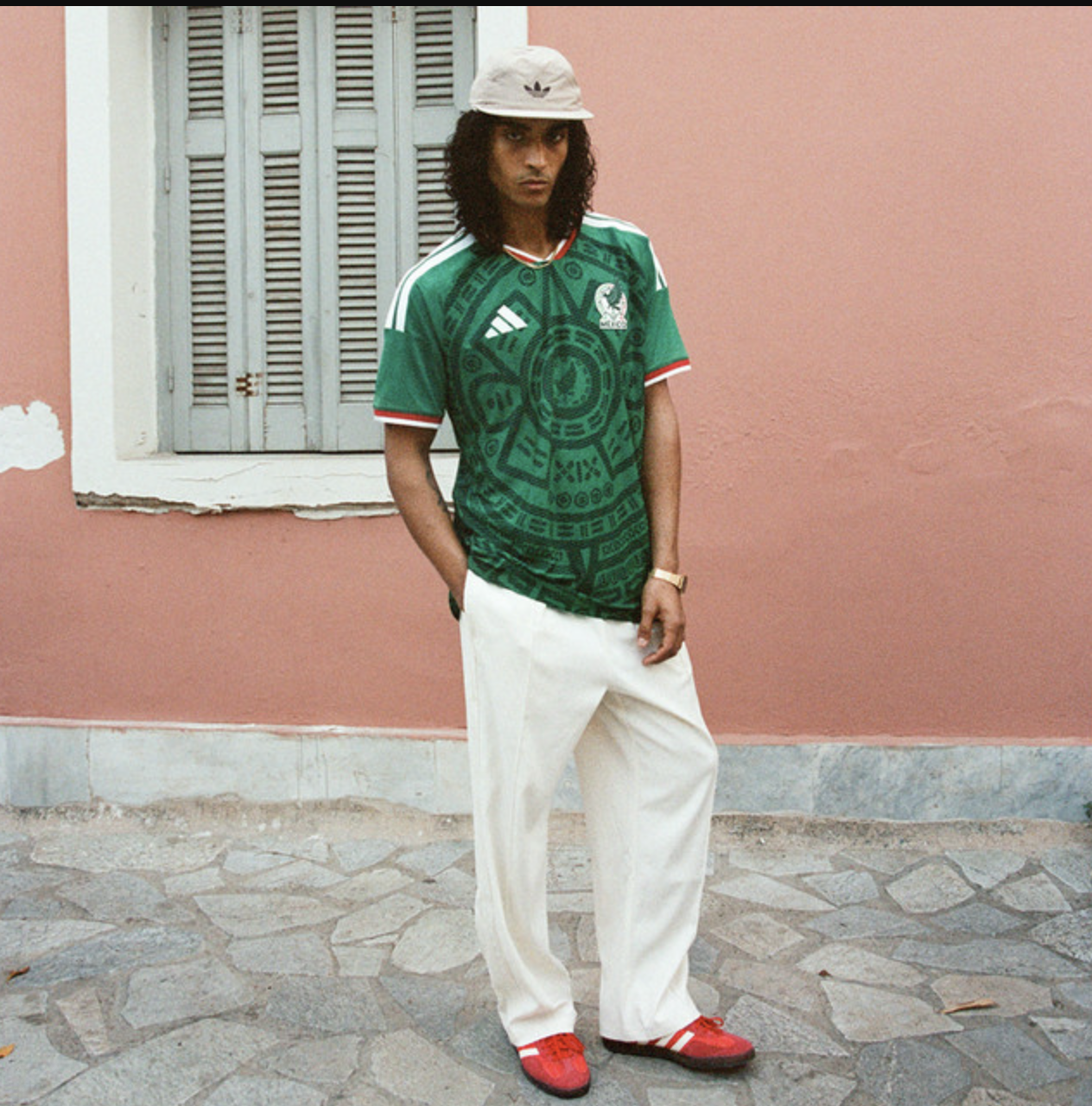 Vibrant streetwear outfit featuring a green graphic Adidas football jersey, wide-leg white trousers, red sneakers, and a beige bucket hat with a black graphic, showcasing contemporary men's casual fashion.